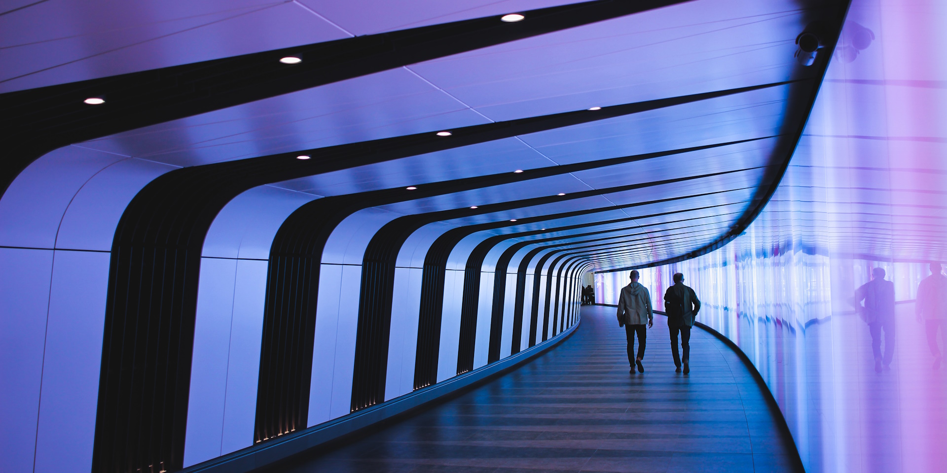 Two men walking in a hallway which has futuristic perspective with purple lighting and reflecting surfaces.