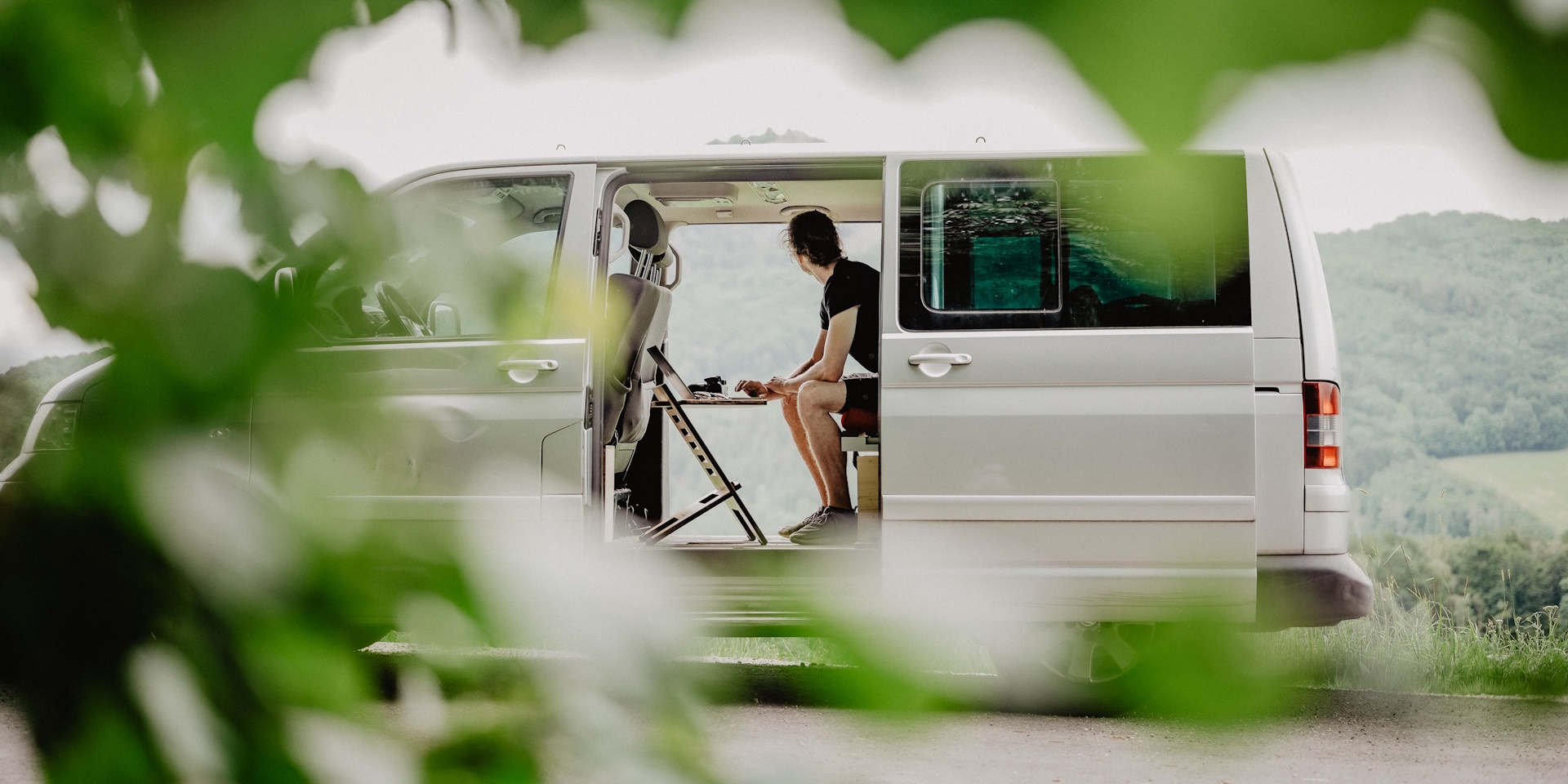 A man working remotely from his van in the middle of nature, using a foldable desk for his laptop
